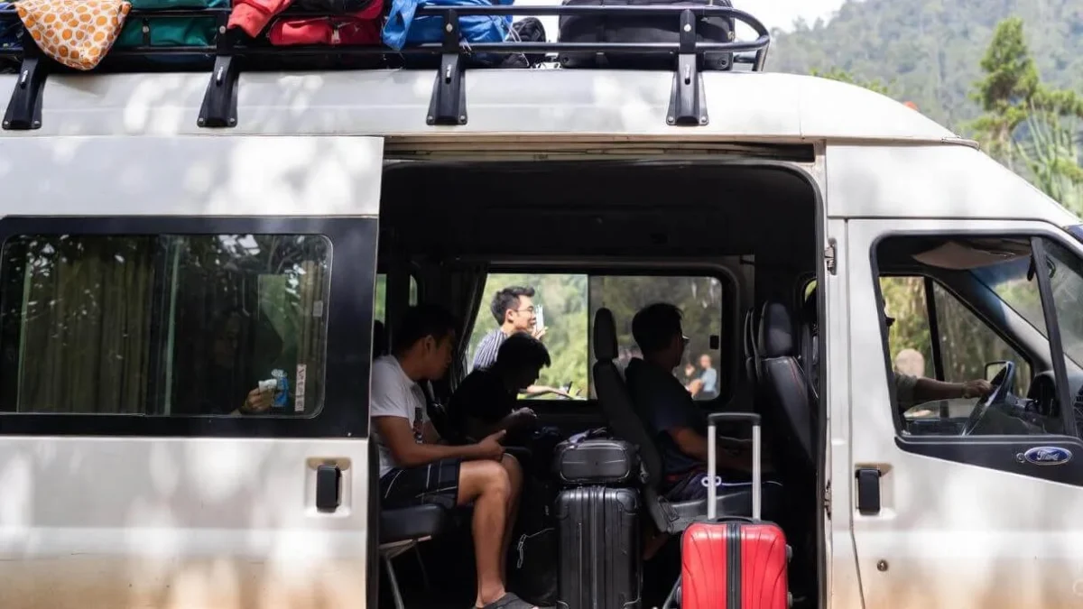 A side view of a white 16-seater passenger van with passengers seated inside and a rooftop rack heavily loaded with colorful luggage.