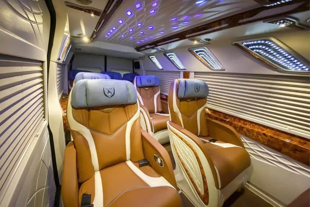 Long-distance van interior with tan and white leather reclining seats and ambient overhead lighting.