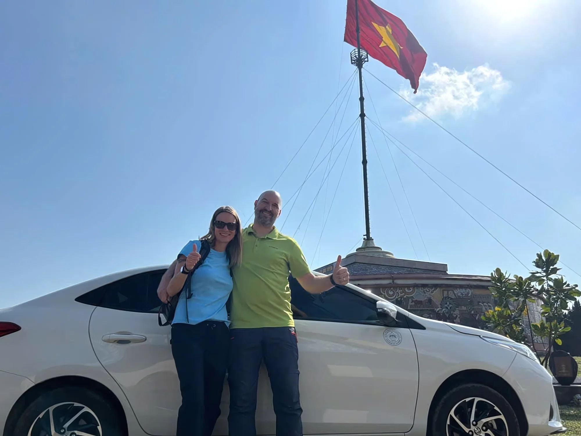 Two travellers giving thumbs up beside a white private car at a Vietnamese landmark