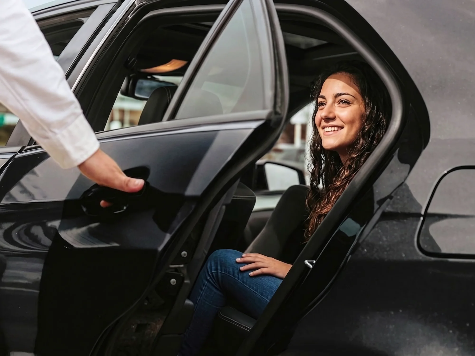 A smiling female traveler seated in a black car backseat as a professional private driver in a white shirt opens the door for a car with driver vietnam service.